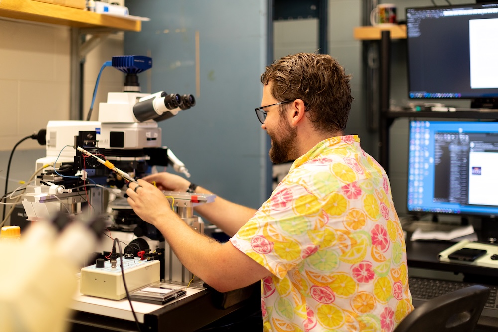 A man in a yellow and pink Hawaiian-style shirt sitting at a microscope